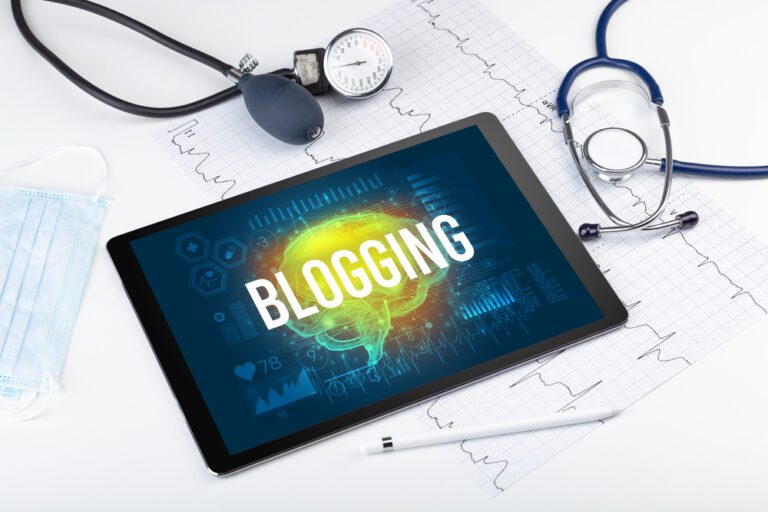 A tablet displaying the word "BLOGGING" with a stethoscope, blood pressure monitor, face mask, and ECG printouts on a white surface—perfect for exploring how to repurpose your healthcare blog content with quick tips for busy providers.