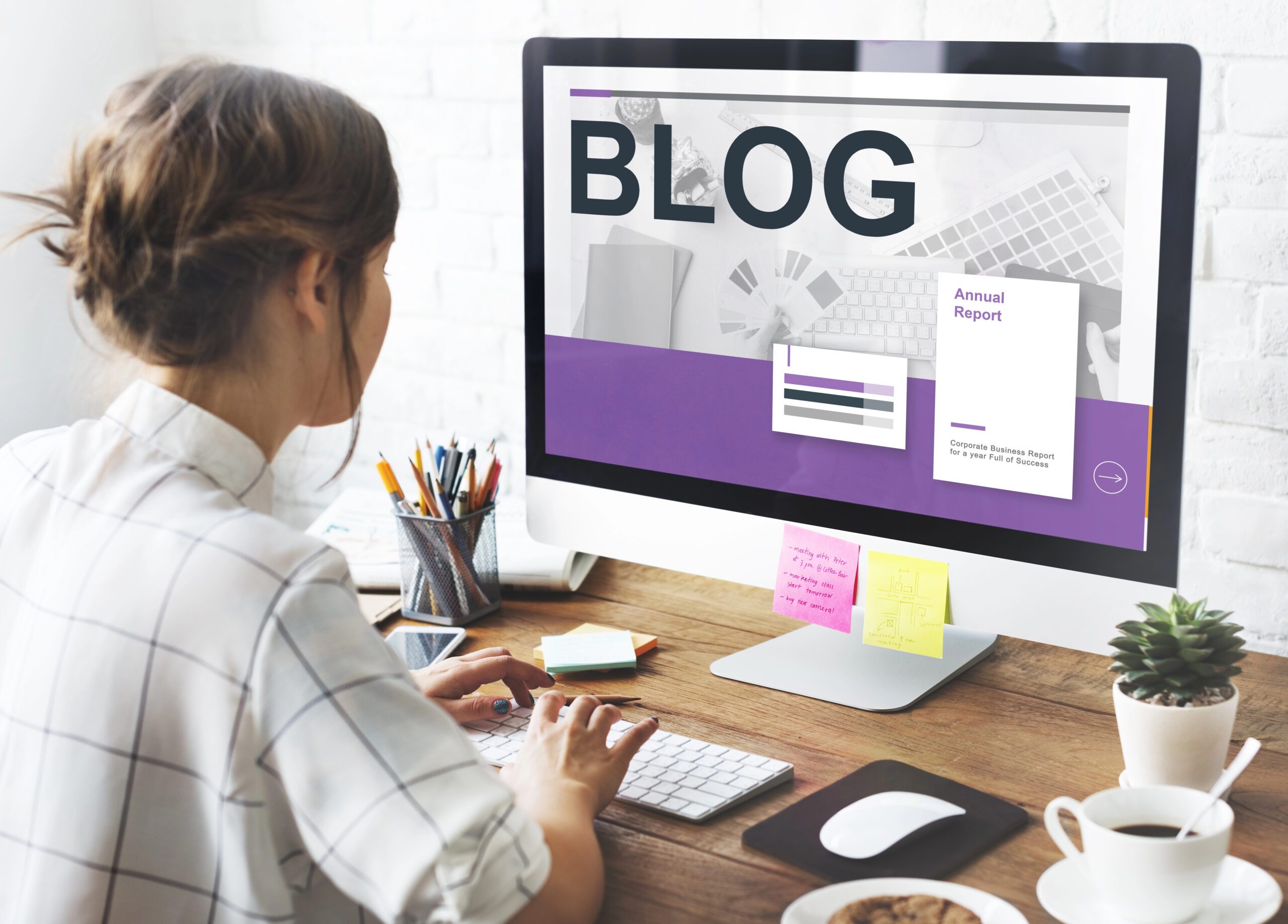 Person typing on a computer with a screen displaying the word 'BLOG' along with a document titled 'Annual Report'. The desk has a cup of coffee, notepads, and sticky notes.