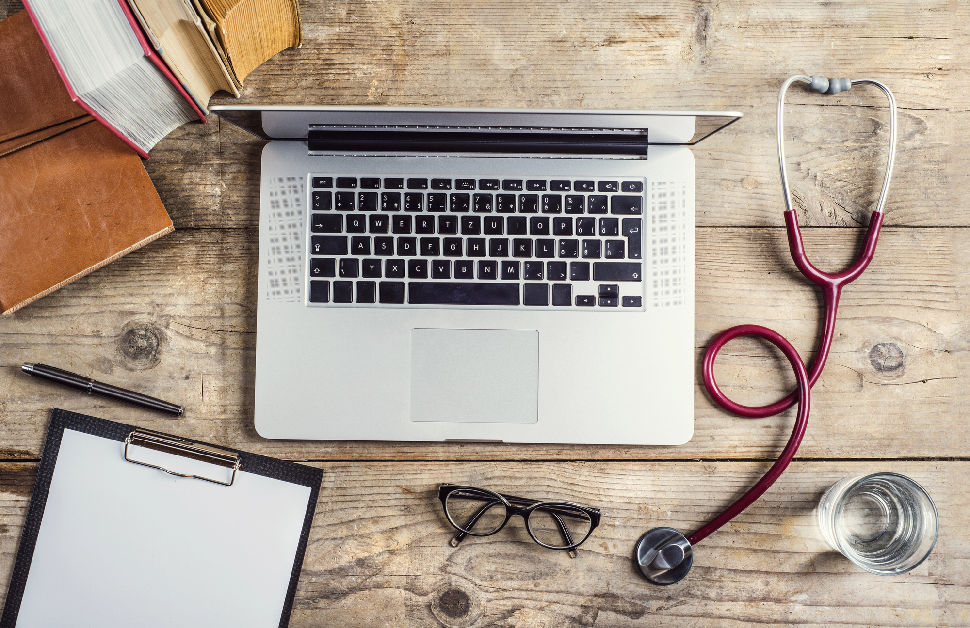 A laptop on a wooden desk surrounded by a stethoscope, books, clipboard, glasses, pen, and a glass of water.