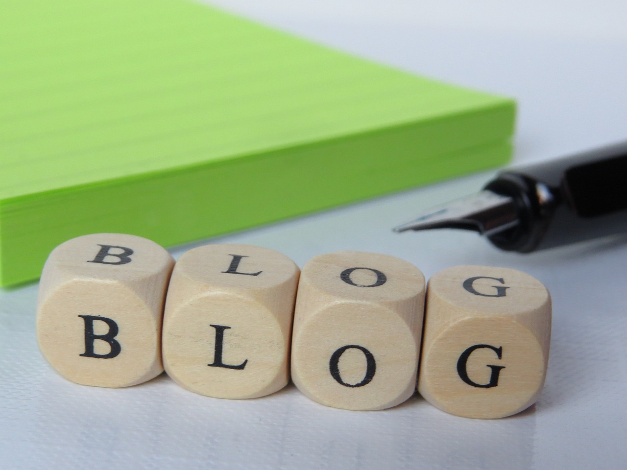 Four wooden blocks spelling "BLOG" are placed in front of a green notepad and a fountain pen.